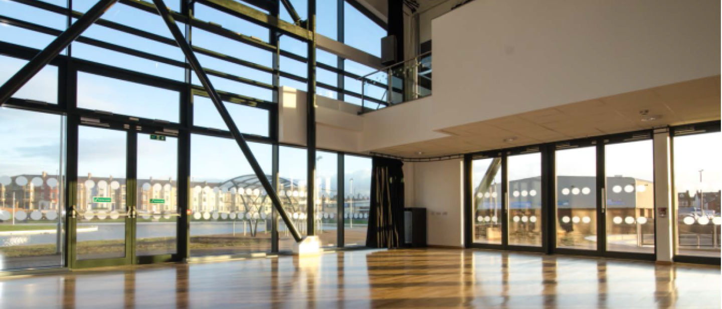 Picture of the main hall in Tuned In, a spacious room with floor to ceiling windows on one of the walls, wooden floor and spotlights on the ceiling.