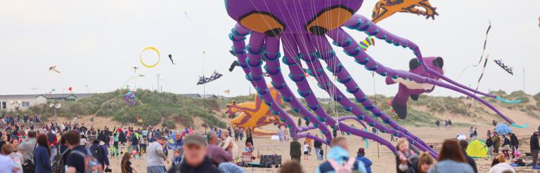 Image from the 2024 kite festival. The image shows a crowd of people on a beach with a purple octopus kite in the background.