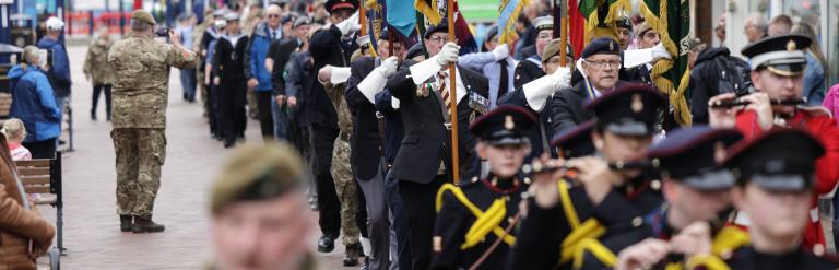 Image of an armed forces parade. A man in camo uniform is in the foreground with marchers in blue outfits in the background with various flags waving.