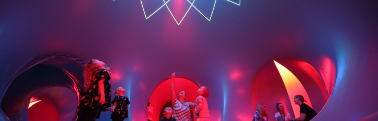 Image of the luminarium as part of between the tides. The image shows families inside a purple and pink room looking at an illumination on the ceiling.