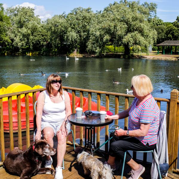 Image of two ladies enjoying the sunshine with their dogs, at the Locke Park café terrace with the lake and trees in the background.