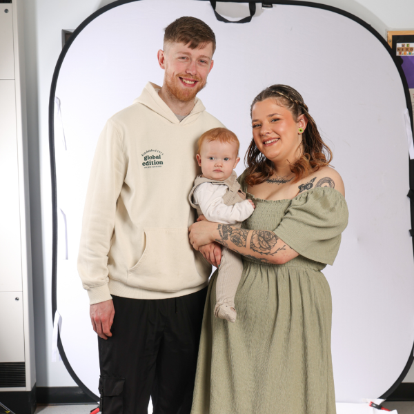 A man and a woman standing in front of the camera and smiling. the woman is holding a baby in her arms.