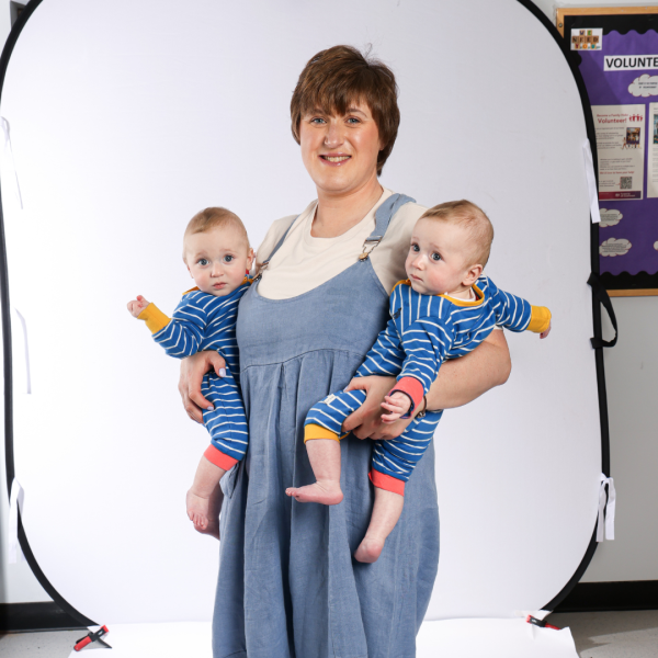 Woman in blue overalls standing in front of the camera and holding two baby boy twins.