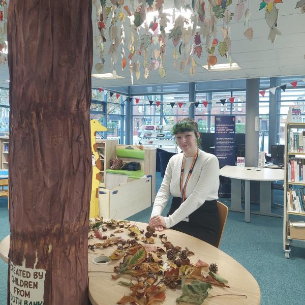 A young girl with green hair wearing a white shirt and black trousers, sitting behind a round table with a decorative real size tree next to her