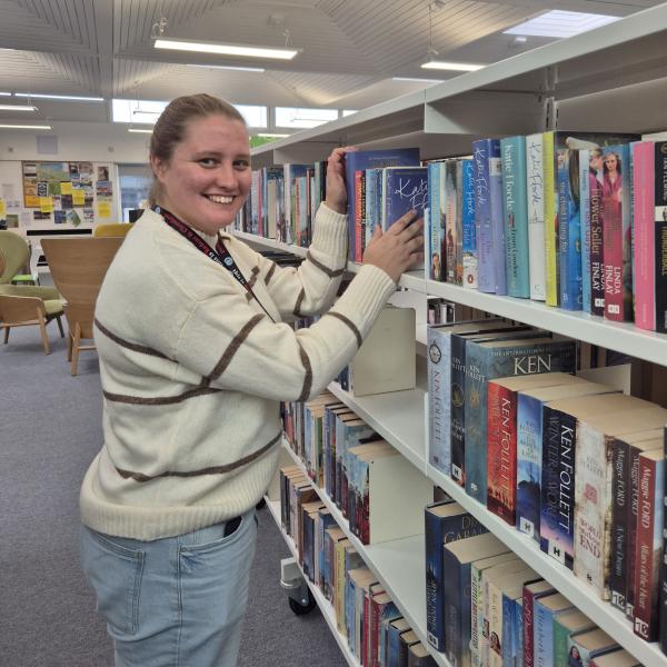 A young woman smiles at the camera while arranging books on a white bookshelf inside a library