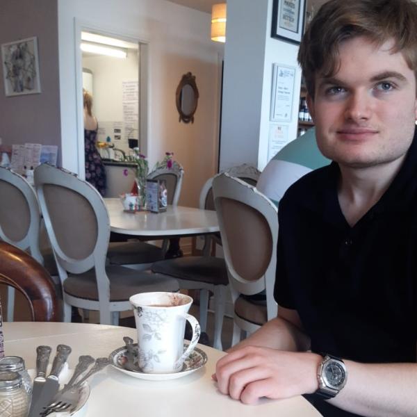 A young man dressed in a black t-shirt sitting at a table in a coffee shop, with a tea cup on the table