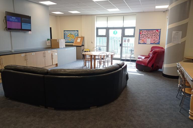 The lounge area at 25K Youth Centre. In the foreground, circular brown couch in front of a TV with a round table surrounded by chairs and two red armchairs in the background and a glass door leading to a terrace.