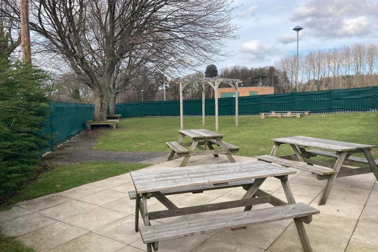 The yard is surrounded by a green fence. Inside the yard, in the foreground, there are three wooden picnic benches on a cement platform, in the background, on a grass covered area there are wooden monkey hanging bars, and two tall tress surrounded by benches